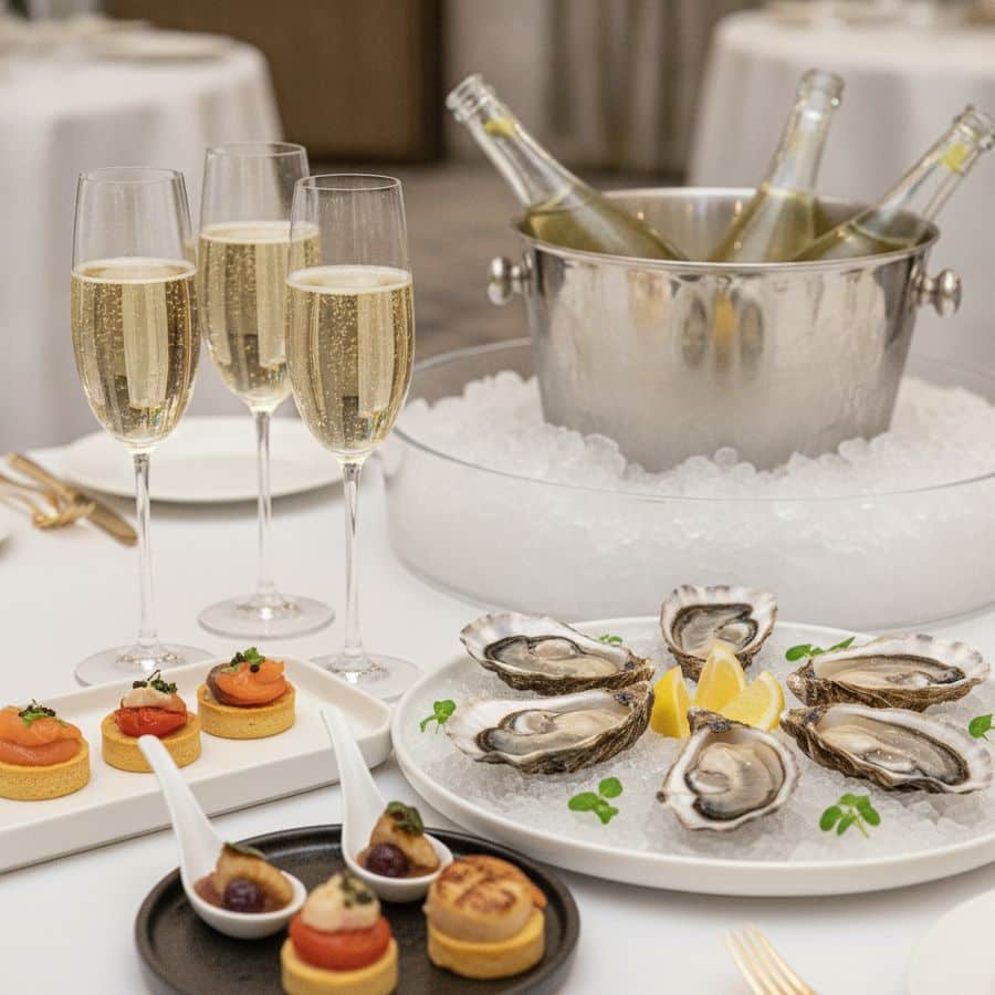 Champagneglazen naast oesters en verfijnde amuses op een chic gedekte receptietafel met heldere belichting.